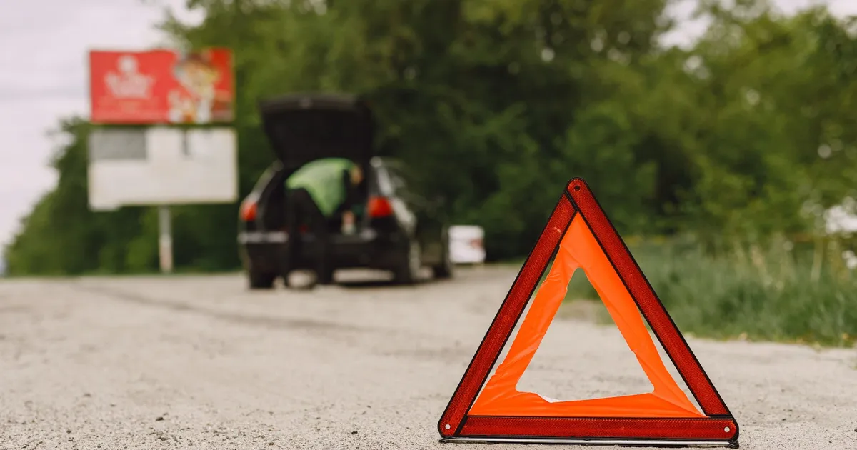 Triangulo de emergencia en carretera con coche averiado de fondo y maletero abierto